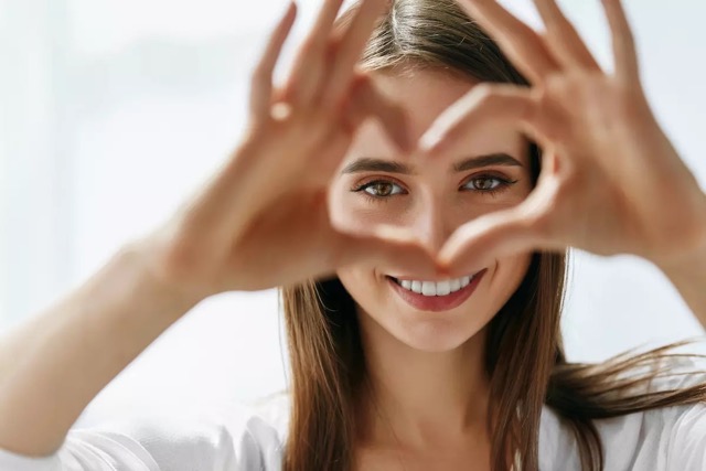 A woman smiles and holds her hands up in a heart shape in front of her eyes.