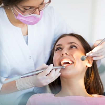 A woman smiles up a dentist or dental hygienist who is placing dental tools in her mouth.
