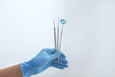 A hand, covered in a blue medical glove, holds up three dental tools, two scraping devices and one mirror.