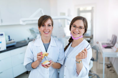 Two young female dentists stand together, holding tools and a model of teeth.