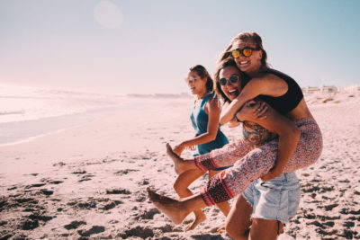 Three young women stand on a beach. One is carrying another woman on her back, piggyback style. They are smiling and wearing sunglasses.