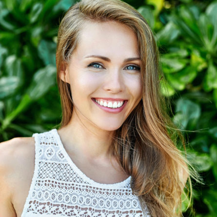 A woman wearing a white crochet top stands in front of a bush. She is in her mid twenties and is smiling.