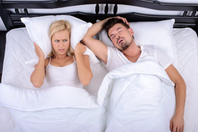 A couple lays in bed, the woman has a pillow held over her ears, and the man appears to be snoring.
