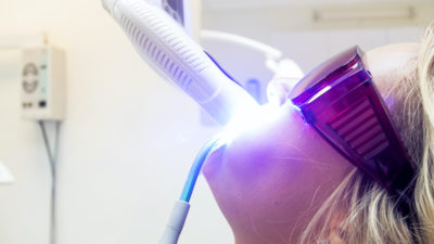 A woman wearing safety goggles receives a tooth whitening treatment with a UV light.