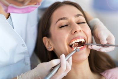 A woman smiles while at the dentist, with a dentist placing tools in her mouth.